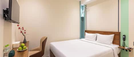 A hotel room with a bed, a desk with a chair, a TV mounted on the wall, and a bedside table with a phone.