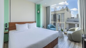 A modern hotel room with a large bed, a seating area with a chair, a small table, and a view of the city through large windows. - V'Luna Hotel Can Tho (Can Tho)