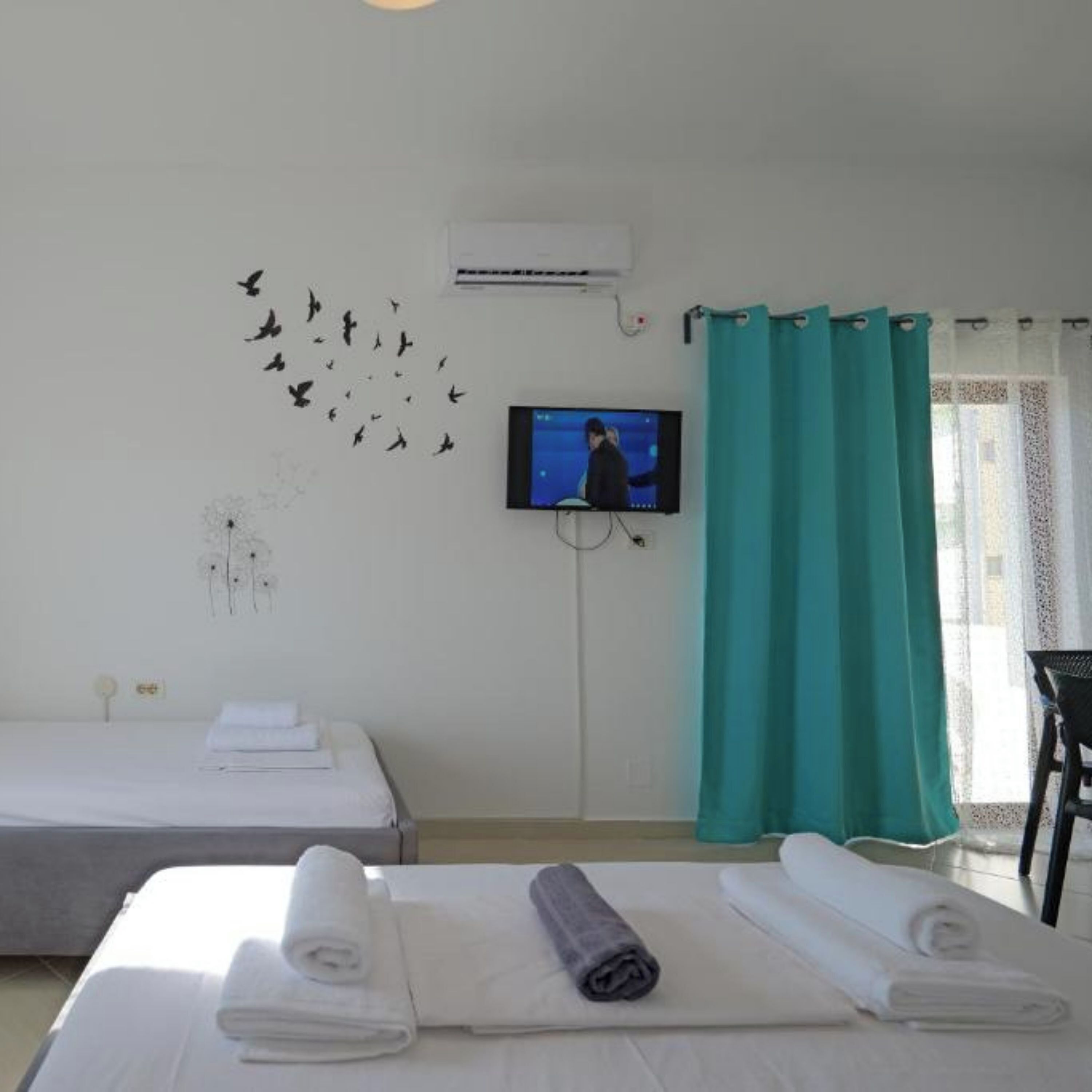 A hotel room with two beds, a television mounted on the wall, and a window with teal curtains.