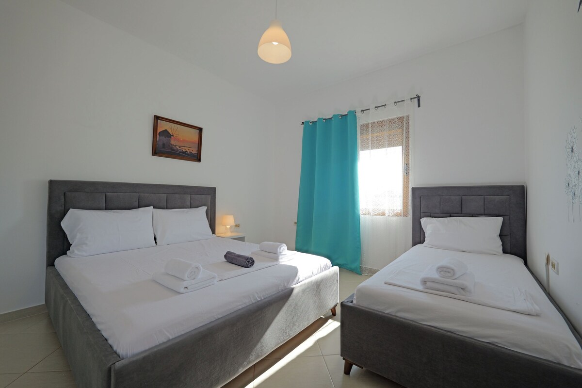 A hotel room with two beds, a painting on the wall, a window with curtains, and a hanging light fixture.