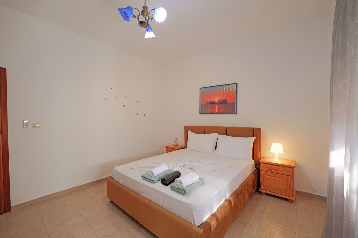 A neatly made bed with white linens and towels, a wooden headboard, a bedside lamp, and a painting of a sunset on the wall.