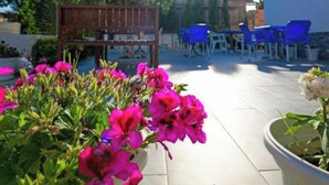 Área de varanda externa com pérgola de madeira, cadeiras de plástico azul e um vaso com flores rosa.