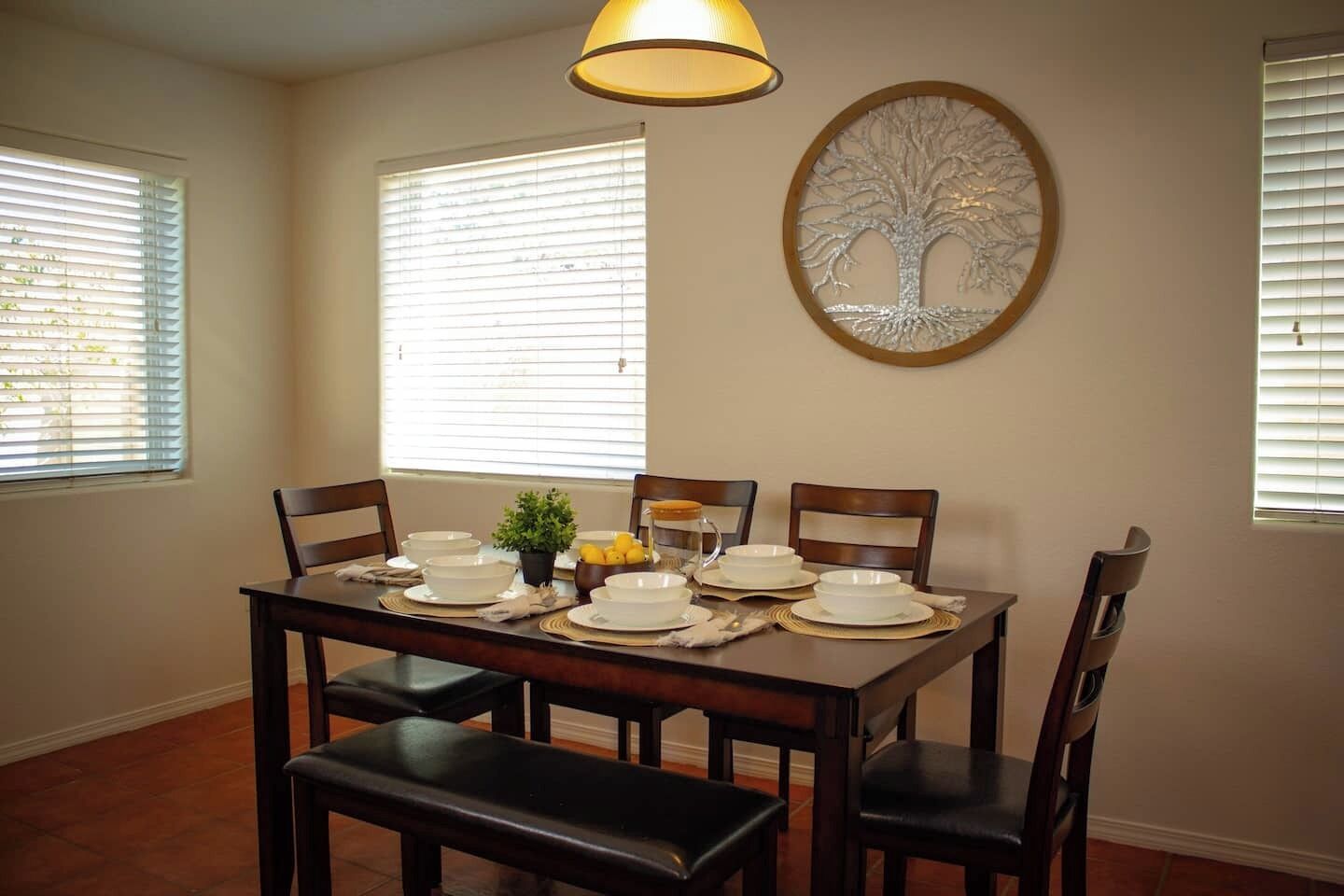 Comedor con una mesa de madera dispuesta para cuatro personas, rodeada de sillas. Hay una planta en maceta sobre la mesa y una obra de arte de un árbol decorativo en la pared.