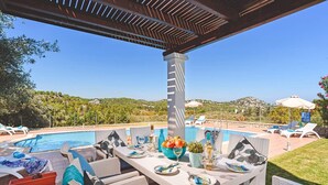 An outdoor pool area with lounge chairs, a dining table set for two, and a view of the surrounding landscape.
