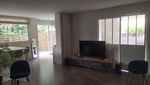 Un salón amplio con un televisor grande, un comedor con sillas y vistas al exterior a través de amplios ventanales.