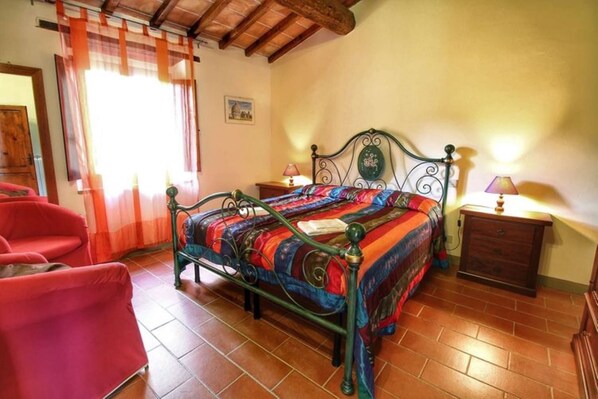 Une chambre à coucher avec un lit en métal, une courtepointe aux couleurs vives, une table de chevet en bois et une chaise rouge.