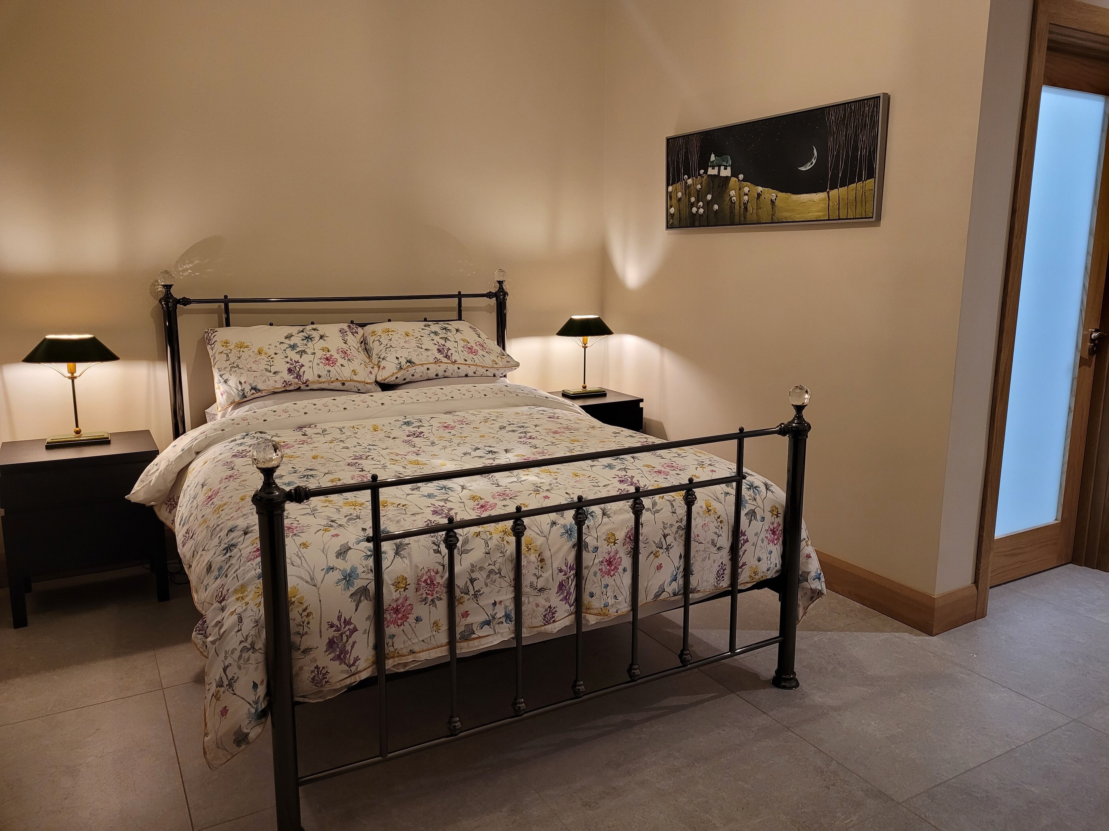 Um quarto com estrutura de cama de metal, roupa de cama florida, duas mesas de cabeceira com abajures e um quadro na parede.