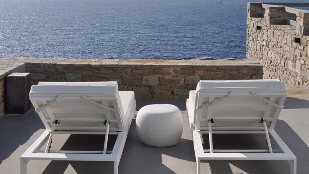 Eine Seeterrasse mit zwei weißen Liegestühlen und einem runden Hocker, mit Blick auf das Meer und vorgelagerte Inseln.