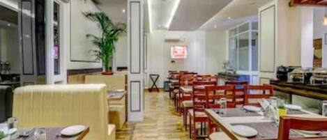 A restaurant with a long counter, multiple tables, and chairs. There is a plant in the corner and a sign on the wall.