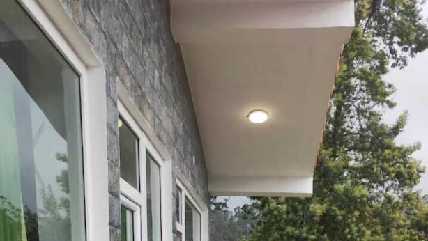 A wooden balcony with a black railing, a stone wall, and a window with a view of trees and houses. - A.V.N Homestay (Kodaikanal)