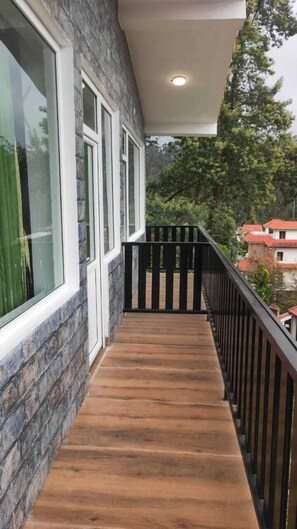 Un balcon en bois avec une balustrade noire, un mur en pierre et une fenêtre donnant sur des arbres et des maisons.