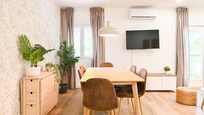 A modern dining area with a wooden table, brown chairs, potted plants, a hanging light, and a flat-screen TV mounted on the wall.