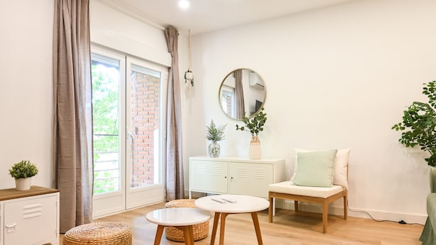 A modern living room with a white sofa, round coffee table, and a small wooden chair. There is a white cabinet with decorative items and a mirror on the wall.