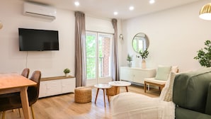 A modern living room with a flat-screen TV, a sofa, a dining table, and a large window with curtains.