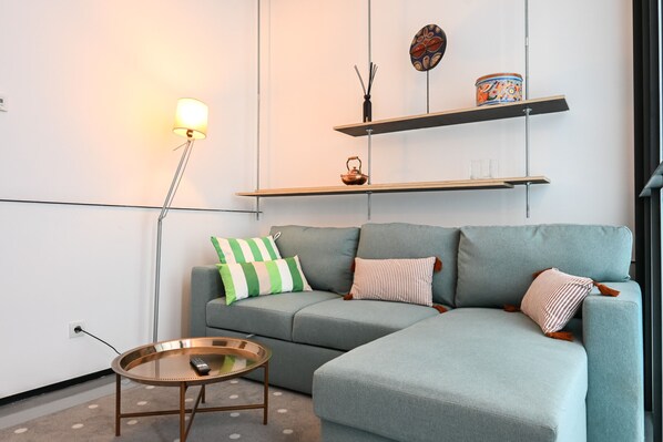 Una sala moderna con un sofá seccional gris, una mesa redonda de centro y una lámpara de pie. Hay estantes sobre el sofá con objetos decorativos.