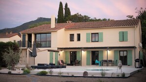 A two-story house with a swimming pool, green shutters, and outdoor seating.