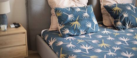 A neatly made bed with a blue and white leaf-patterned bedspread, a bedside table with a lamp and remote controls, and a grey headboard.
