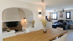 A modern living room with a fireplace, white sofas, a wooden dining table, and a ceiling fan.
