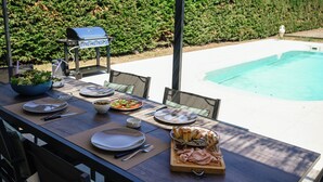 A dining table set for a meal with plates, cutlery, and a variety of food items, positioned outdoors with a pool and greenery in the background.