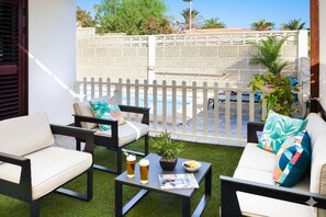 An outdoor seating area with two sofas, a coffee table with drinks and a plant, and a view of a pool and greenery.