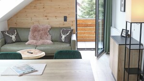 A cozy room with a wooden ceiling, a green sofa with deer-themed cushions, a fluffy pink throw, a wooden coffee table, and a small table with a lamp and picture frame.