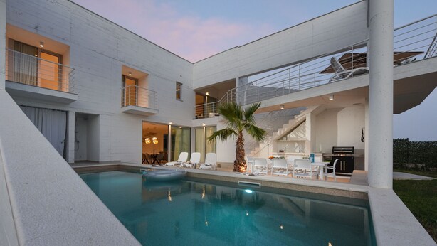 Ein modernes Haus mit Schwimmbad, Sitzgelegenheiten im Freien und einer Treppe, die zu einem Balkon führt.