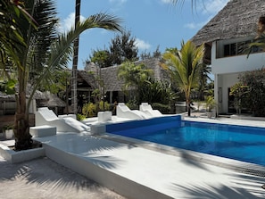 A pool area with white lounge chairs, a clear blue pool, and thatched-roof structures. - Ayras Hotel Zanzibar (Paje)