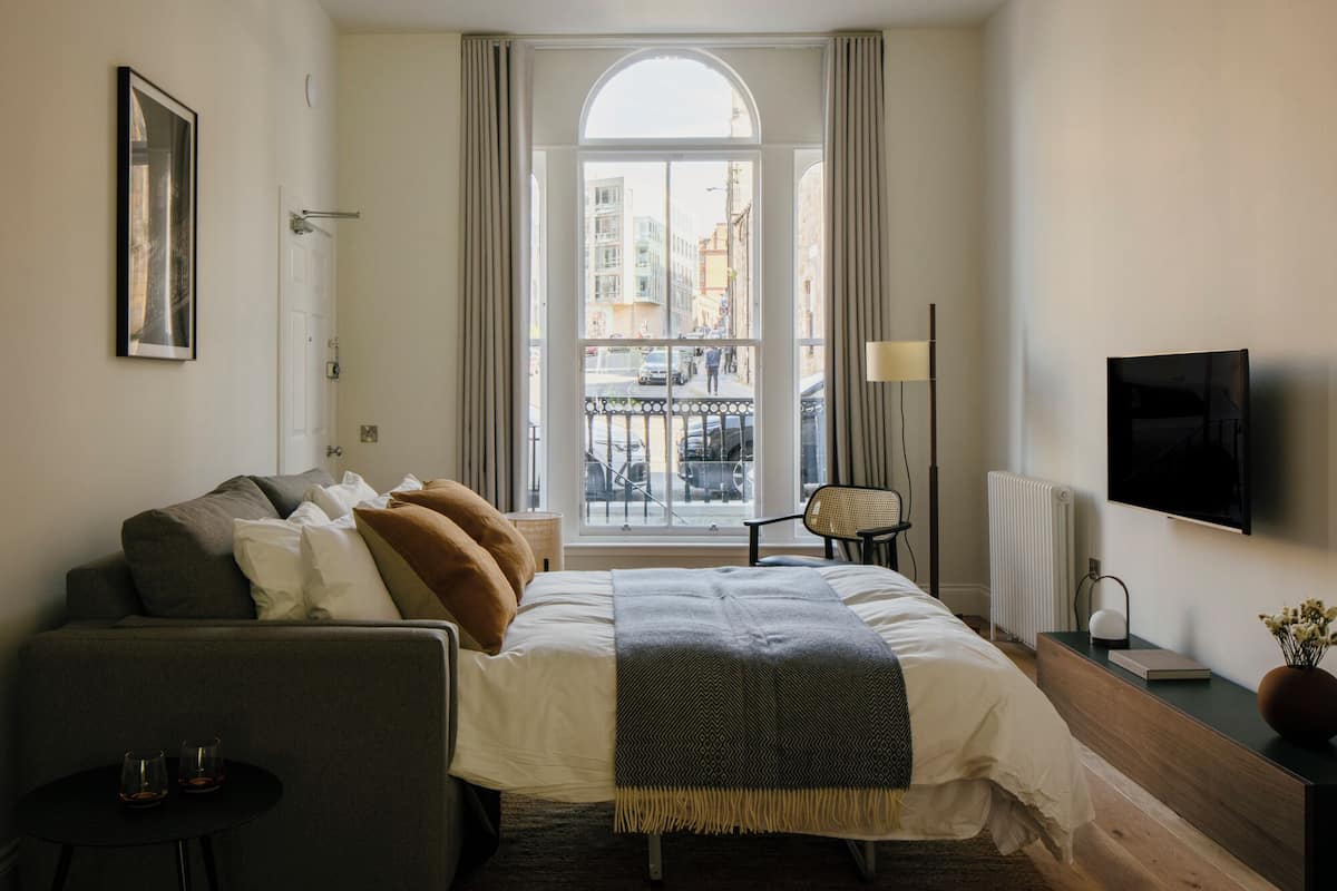 A modern hotel room with a large bed, a sofa, a small table, a chair, a TV, and a window with a view of the city.