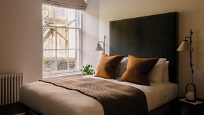 A neatly made bed with a dark headboard, two bedside lamps, and a window with a view of a stone building. - 33 Castle Terrace (Edinburgh)
