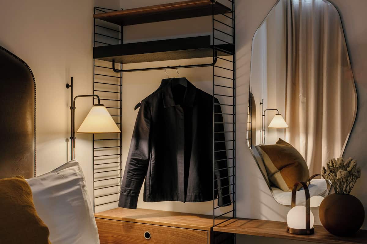 A bedroom with a bed, a wooden nightstand, a mirror, a hanging coat rack, and a lamp.