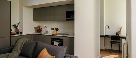 Un apartamento moderno con un sofá gris, una pequeña cocina con armarios oscuros y una zona de comedor con una silla y mesa de madera.