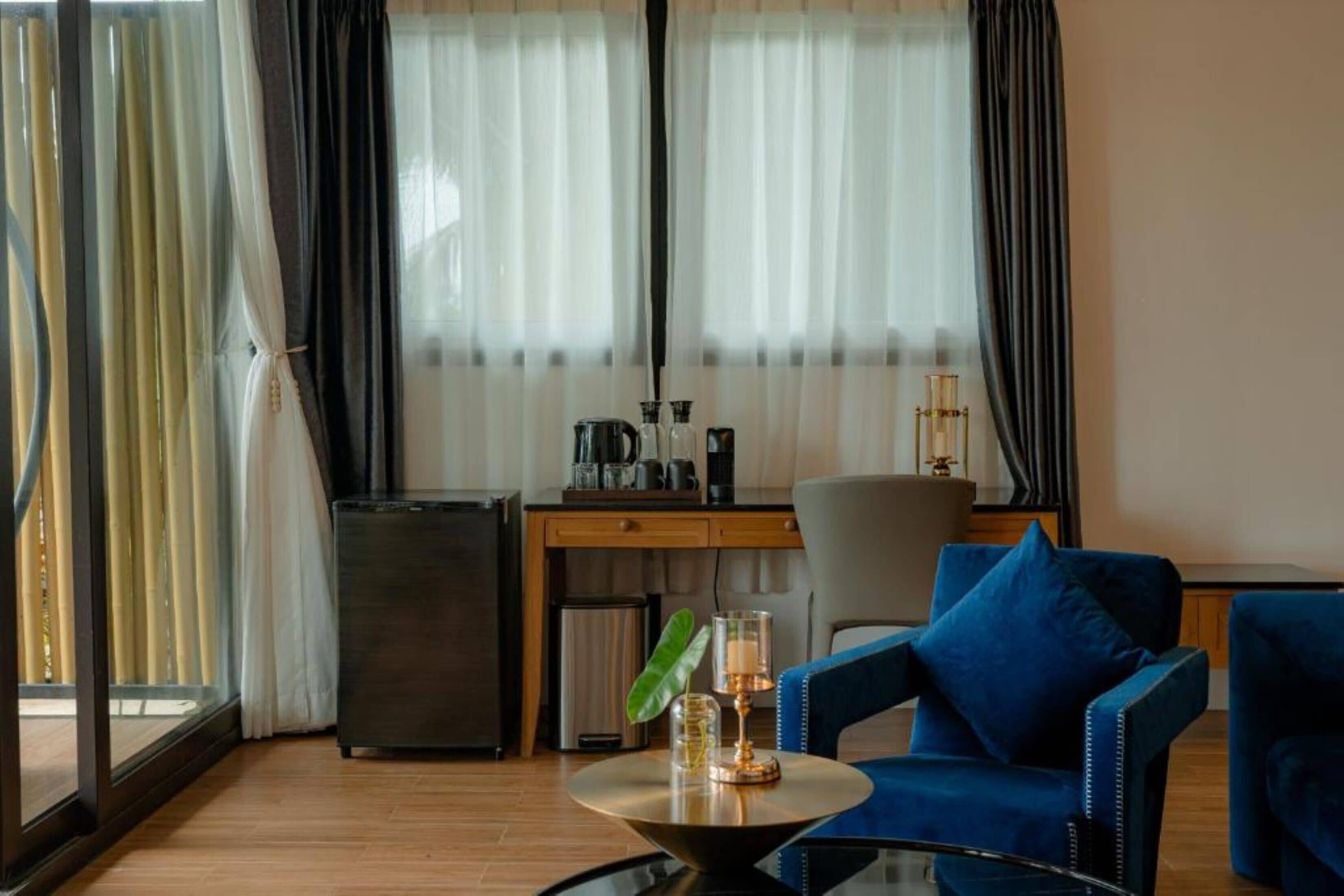 A modern hotel room with a wooden floor, a blue sofa, a small table with a lamp, a desk with a chair, and a minibar.