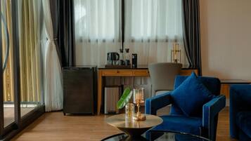 A modern hotel room with a wooden floor, a blue sofa, a small table with a lamp, a desk with a chair, and a minibar.