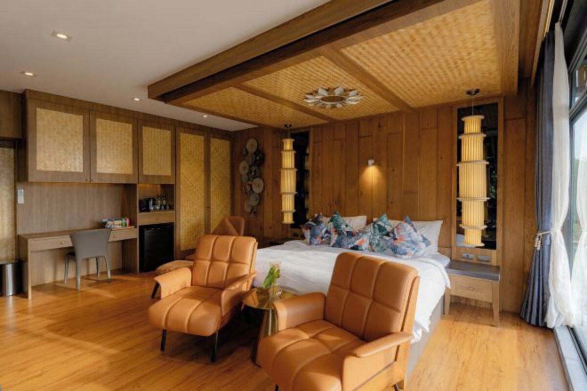 Habitación de hotel moderna con techo de madera, una cama grande con muchas almohadas, dos sillones de cuero, un escritorio pequeño y un ventanal con cortinas.