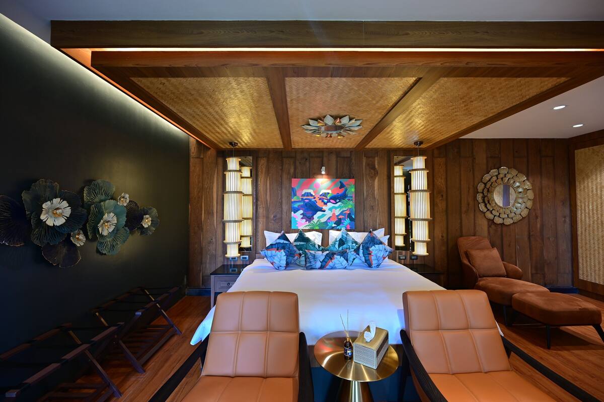 A modern hotel room with a large bed, two brown leather chairs, a small table with a lamp, and a decorative wall piece.