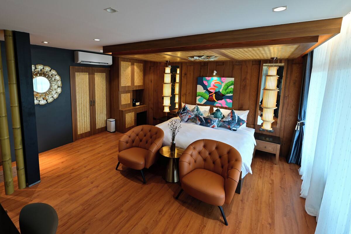 A modern hotel room with a wooden floor, a large bed with a colorful artwork, two leather armchairs, and a small round table.