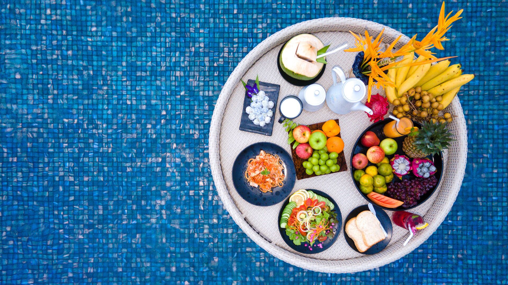 A round tray with various food items and drinks, including fruits, vegetables, and a salad, placed on a blue textured surface.
