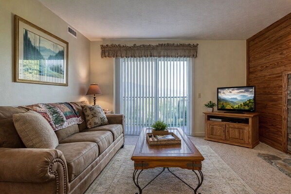 A living room with a sofa, coffee table, TV stand, and a large window with blinds.