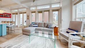 A spacious living area with a glass coffee table, a sofa, and a chair. There is a dining area with chairs and a table. The room has large windows and a ceiling fan. - Resort Pools, Golf Cart Boardwalk to the Beach (Port Aransas)