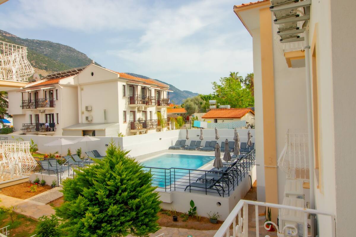 Un espace piscine doté de chaises longues et de parasols, entouré de bâtiments avec des balcons et surplombant un paysage montagneux.