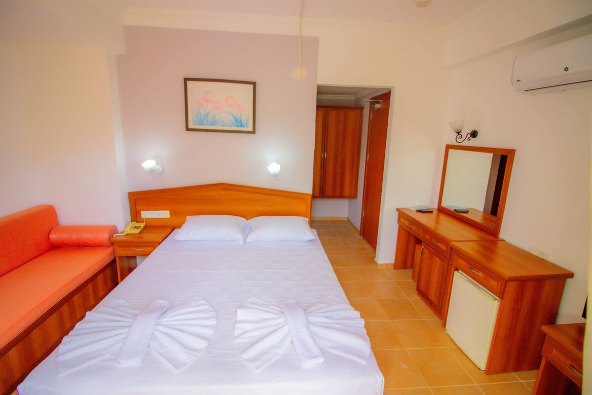 Une chambre d’hôtel avec un lit, une table de chevet en bois, un bureau avec un miroir, un petit réfrigérateur et un canapé rouge.