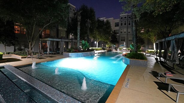 Uma área de piscina noturna com várias fontes, espreguiçadeiras e ambiente bem iluminado.