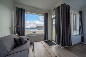 Una habitación con un sofá gris, una mesa de madera y vistas a una playa y montañas a través de amplios ventanales.