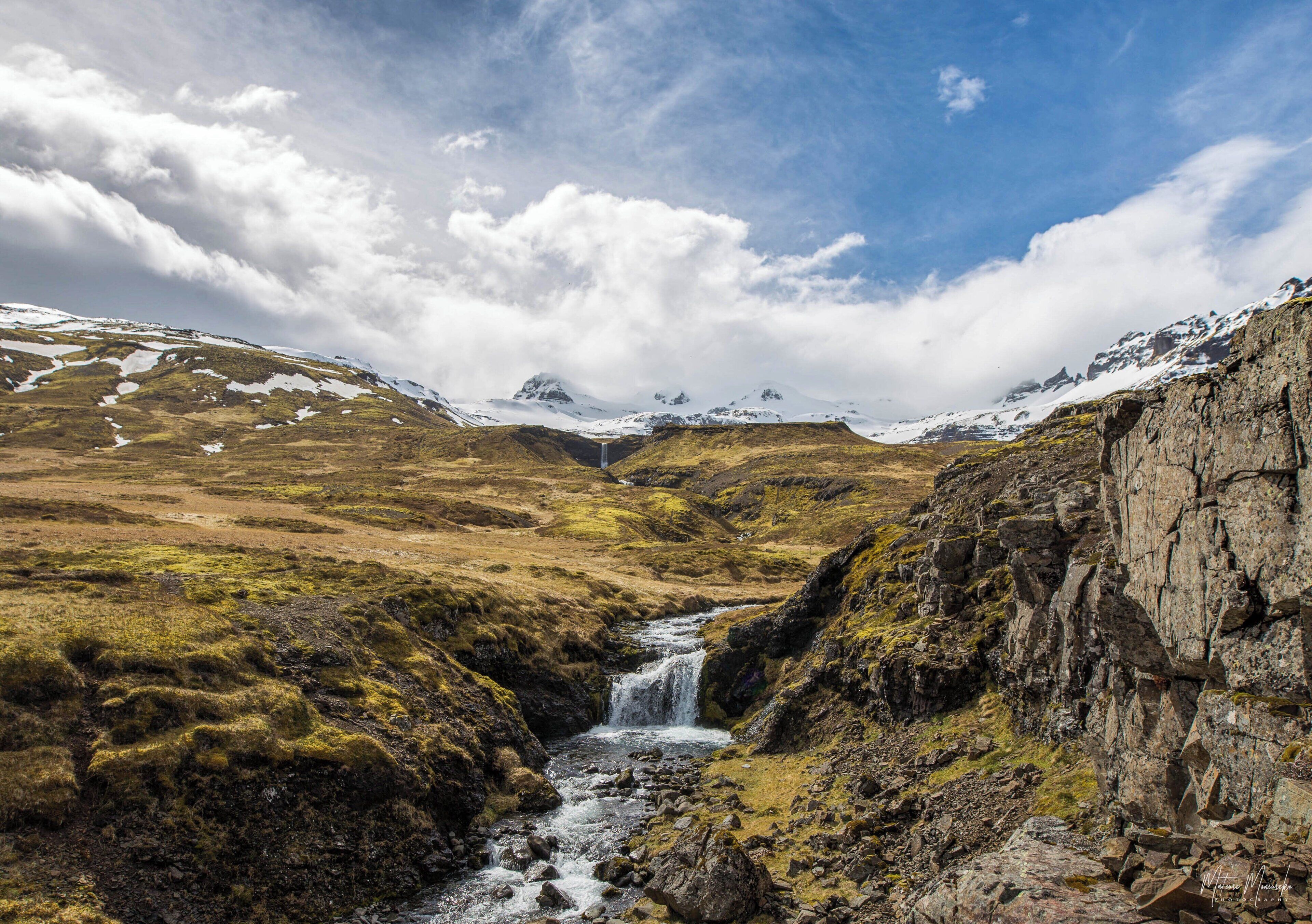 A mountainous landscape with a waterfall, rocky terrain, and snow-covered peaks.