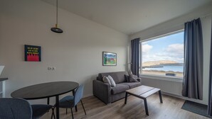 A modern living room with a sofa, a coffee table, and a dining table with chairs. There is a large window with curtains, a hanging light fixture, and two framed pictures on the wall.