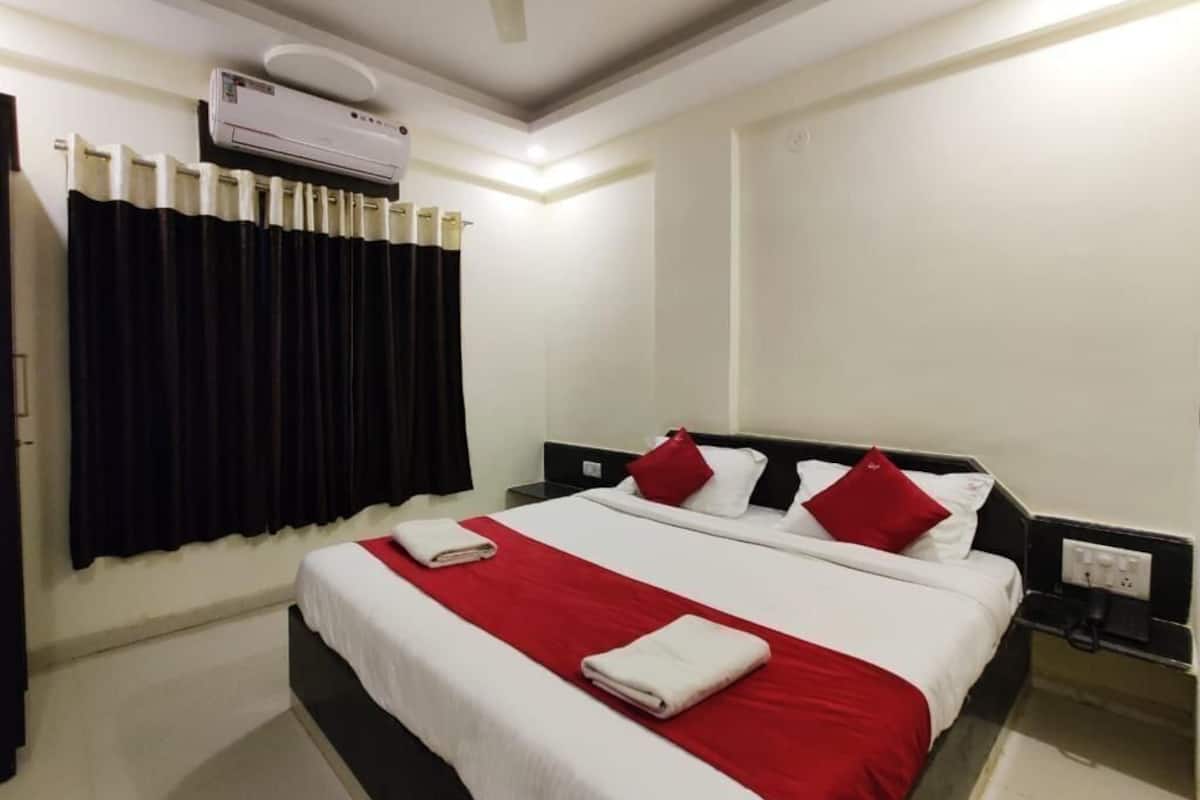 A hotel room with a bed, a ceiling fan, an air conditioning unit, a window with curtains, and a bedside table with a lamp.