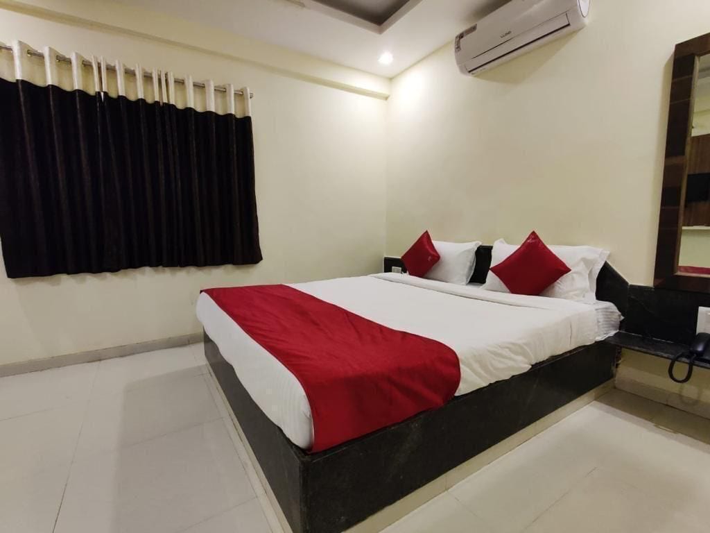 A hotel room with a bed, a black and white headboard, a red and white bedspread, a black curtain, a white air conditioning unit, and a mirror on the wall.