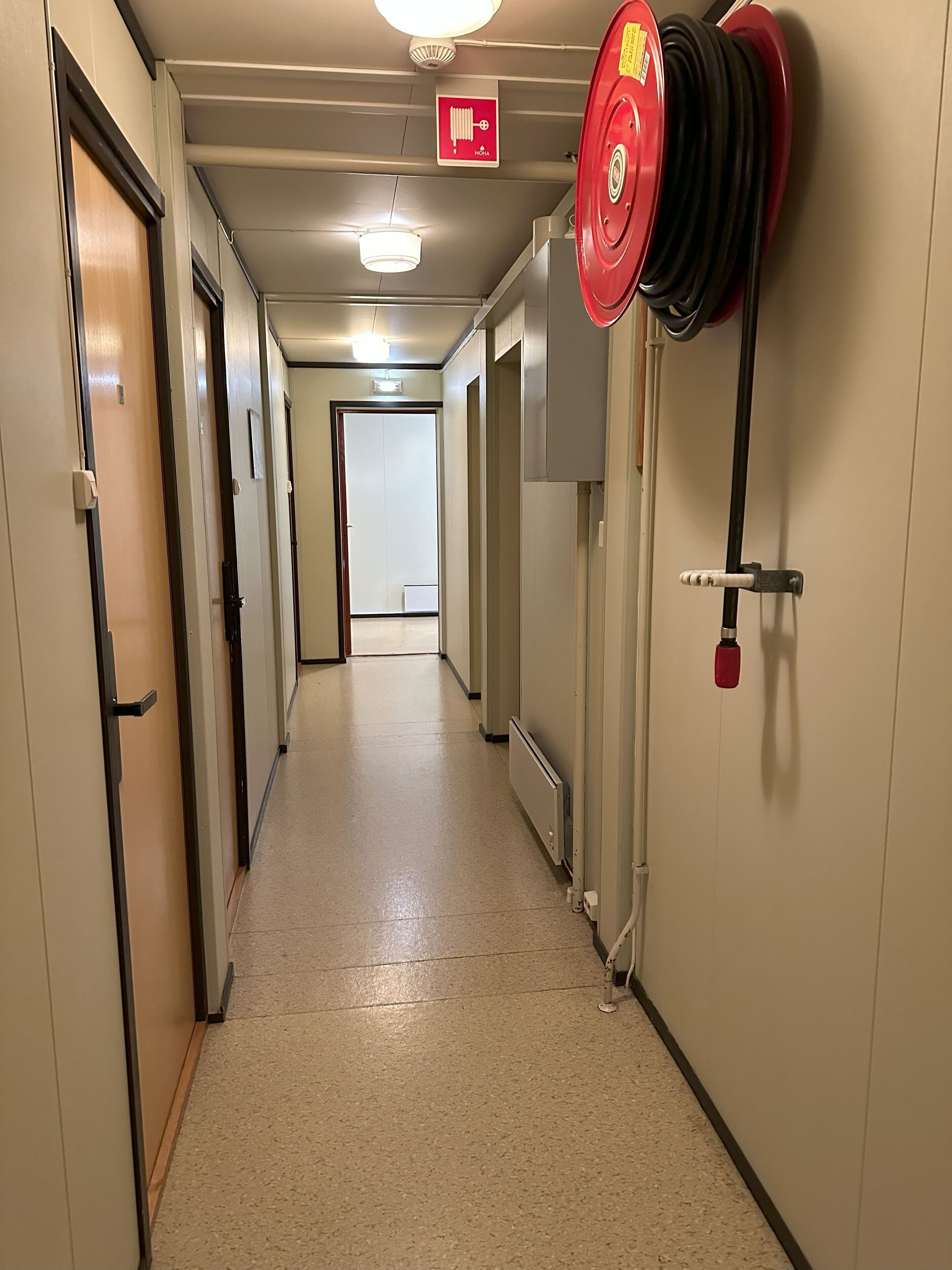 En hotellkorridor med dörrar på båda sidor, en skylt som visar var brandlarmet finns och en röd brandslangsvinda på den högra väggen.