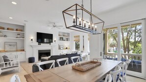 A modern living room with a fireplace, a dining area, and a large window with a view of trees. - 7 BR 8 BA Private Pool Singleton Beach Sleeps 24 Bikes & Beach Gear included (Hilton Head Island)