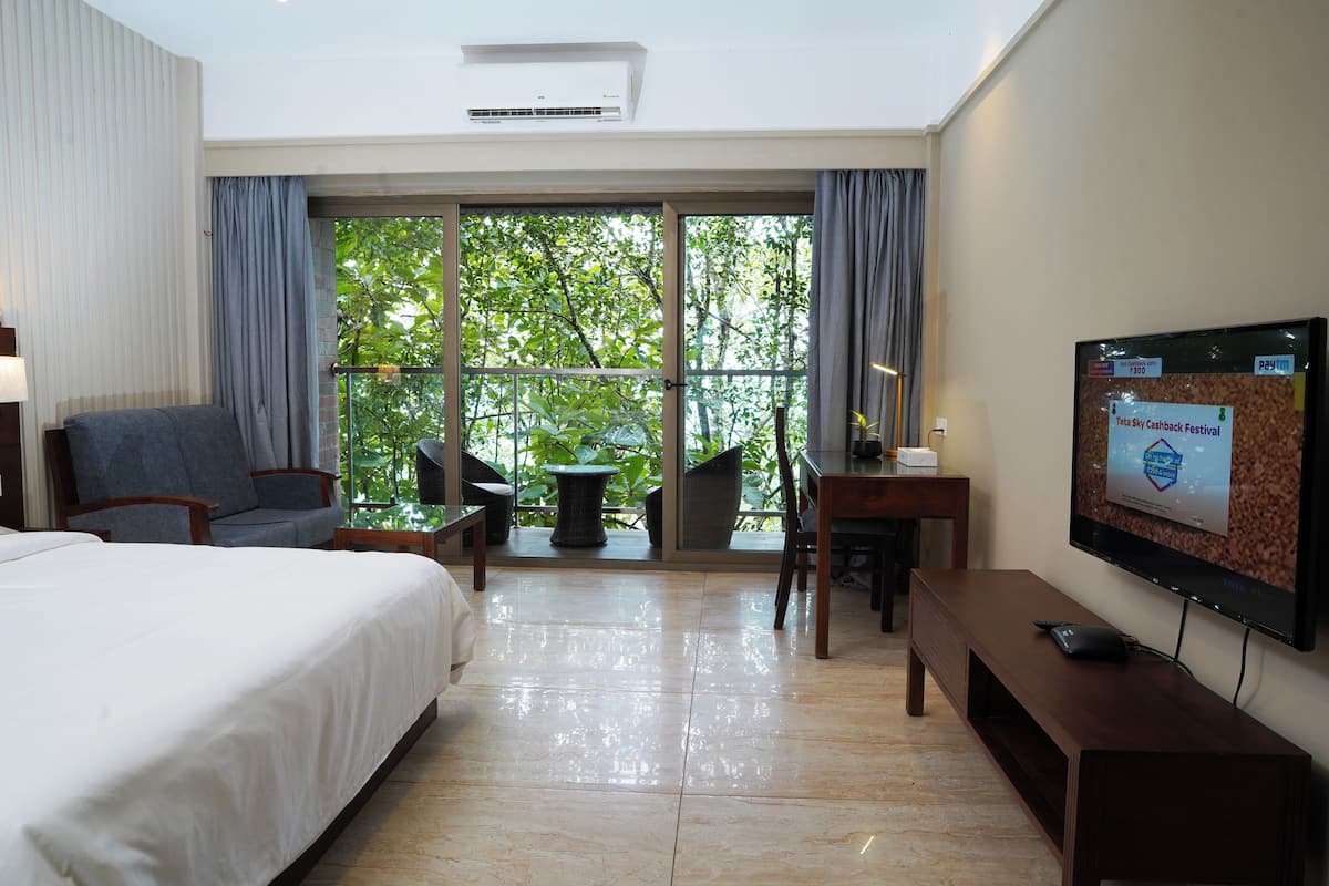 A hotel room with a bed, a desk, a chair, a TV, and a balcony with a view of trees.